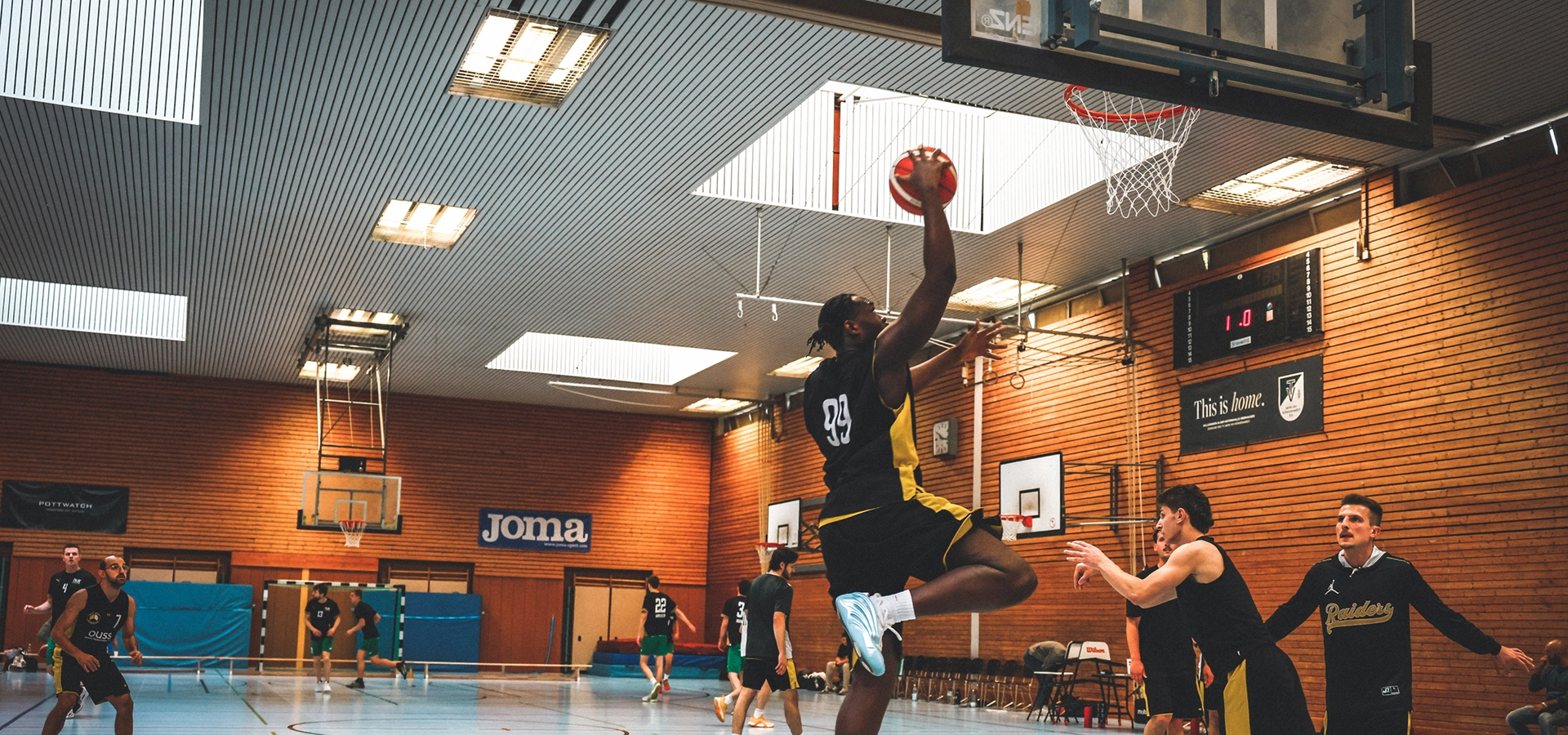 Ein Basketballspieler der SpVg 0507 Odenkirchen in einem schwarzen Trikot springt, um einen Aufschlag zu machen, während die Verteidiger versuchen, ihn in einer Turnhalle zu blockieren. Andere Spieler und ein Trainer beobachten das Geschehen vom Spielfeld aus.