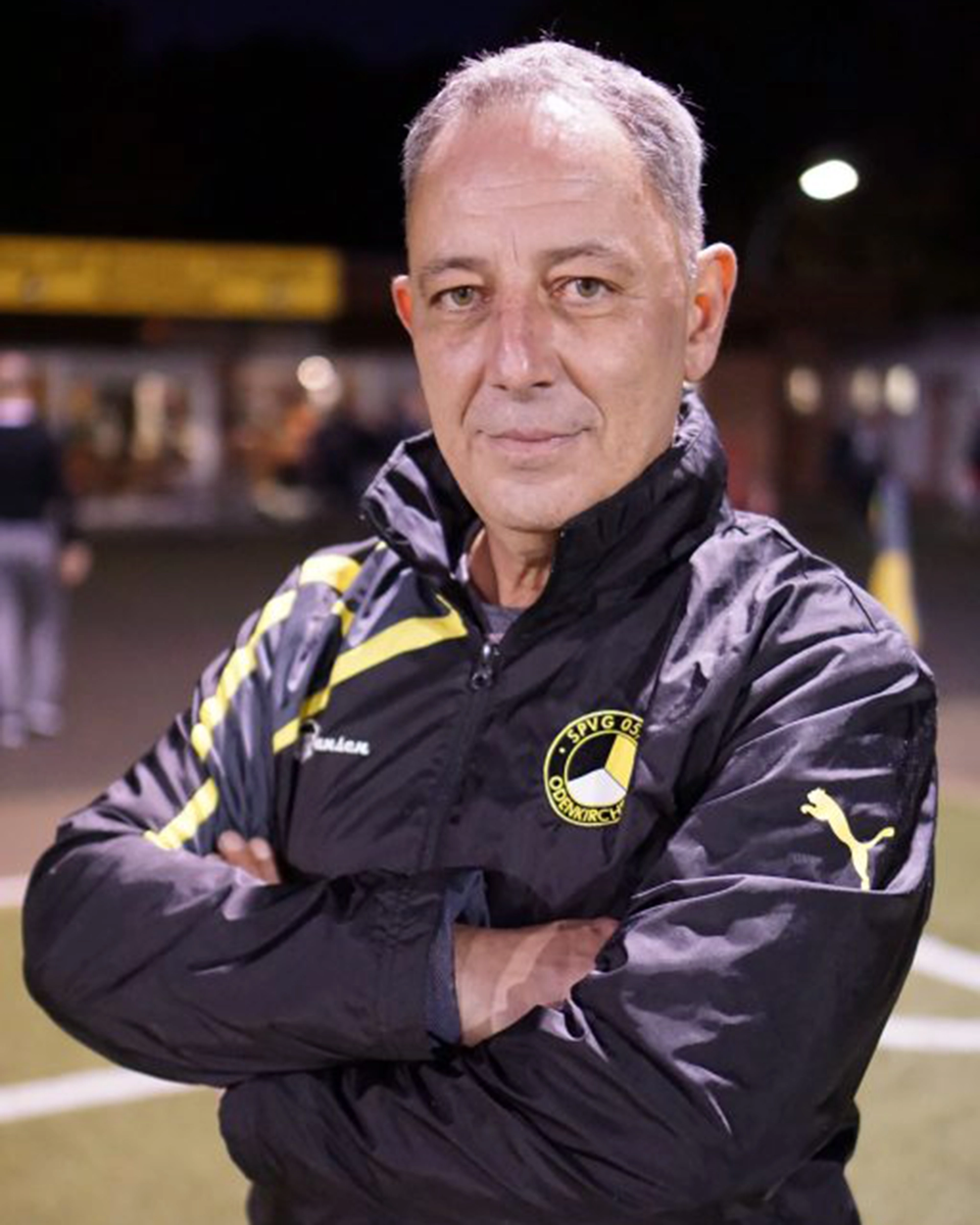 Ein Mann mit kurzen grauen Haaren, der eine schwarze Sportjacke mit gelben Details trägt, steht mit verschränkten Armen auf einem Sportplatz und blickt in die Kamera - ideal für ein Profil des Präsidiums Kontakt. Der Hintergrund ist leicht unscharf und zeigt Lichter und Gebäude.