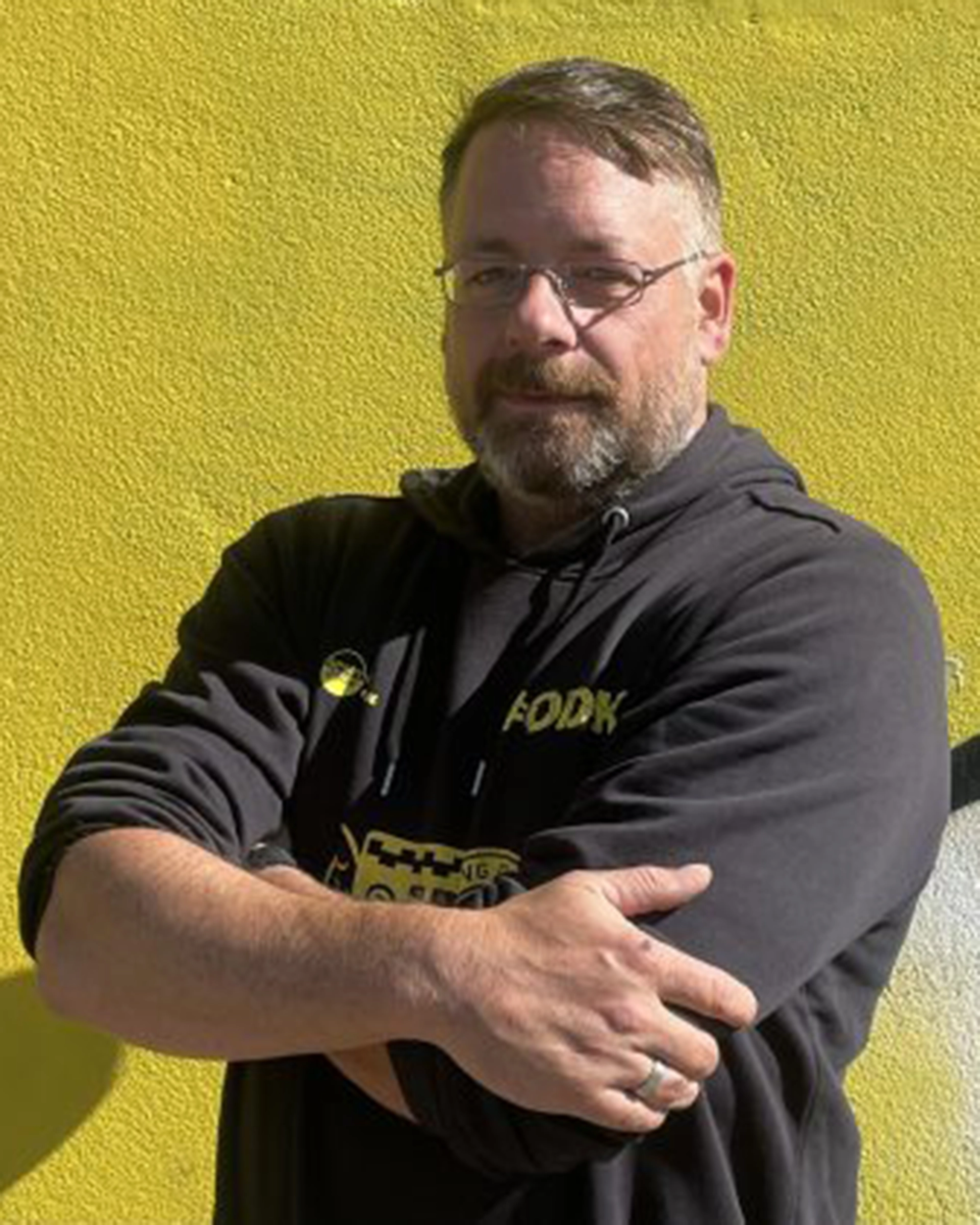 Ein Mann mit Brille und Bart steht vor einer leuchtend gelben Wand und trägt einen schwarzen Kapuzenpulli mit gelber Aufschrift. Mit verschränkten Armen blickt er in die Kamera und verkörpert damit das selbstbewusste und zugängliche Auftreten des Präsidium Kontakt-Teams.